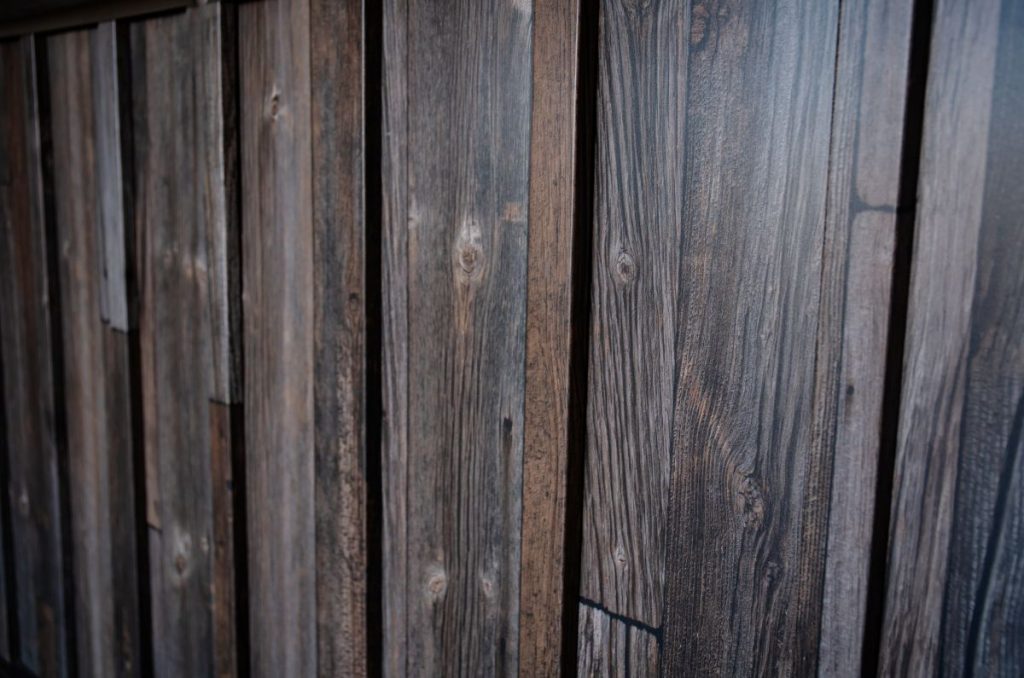 Close-up of a wooden fence with vertical planks featuring a natural, weathered texture in shades of brown and gray. The wood grain is prominently visible, giving the fence an aged and rustic appearance.