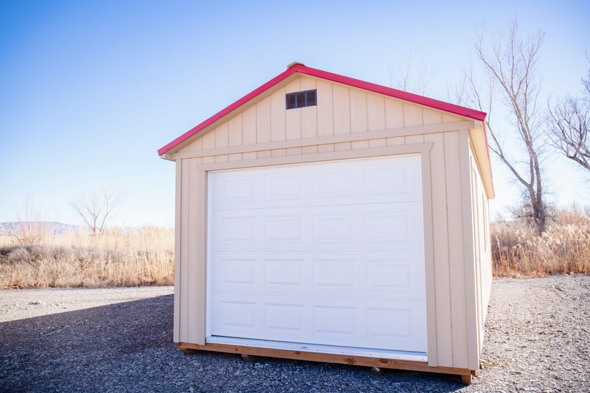 process-step-2-1 A small, beige shed with a white garage door, a red roof, and a small vent at the top, stands on a gravel surface. In the background, leafless trees and dry vegetation are visible under a clear blue sky.