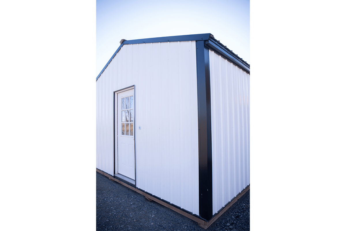 process-step-2-1 A small white metal shed with black trim is shown from an angled perspective. It has a single white door with a window panel. The bottom of the shed is surrounded by gravel. The sky is clear in the background.