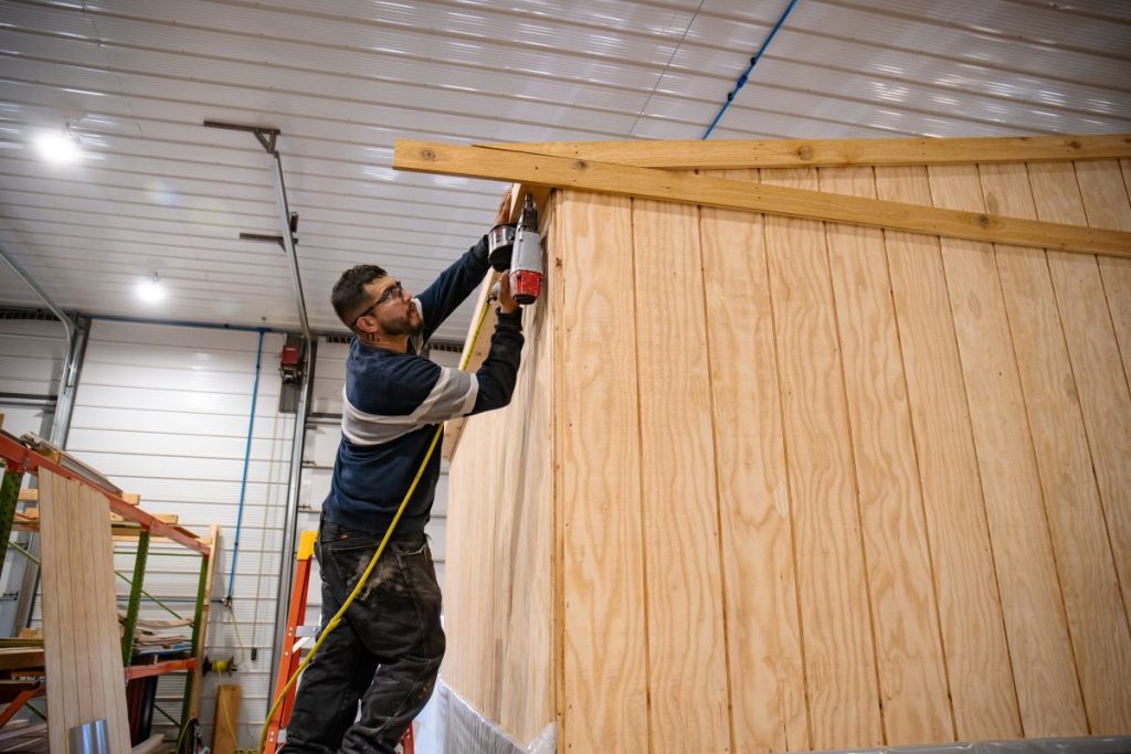 A person in glasses uses a nail gun to attach wood panels to the slanted roof of a wooden structure inside a large, well-lit workshop. The person is wearing a striped sweater and black pants. Nearby, more wooden panels and construction materials are visible.