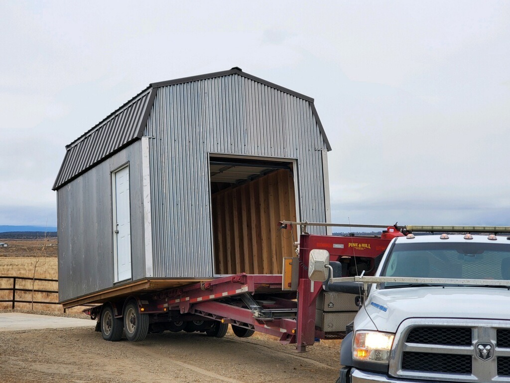 A silver shed on a red trailer is being transported by a white truck on a dirt road. The shed is elevated at an angle on the trailer. The background features a rural landscape with a cloudy sky.