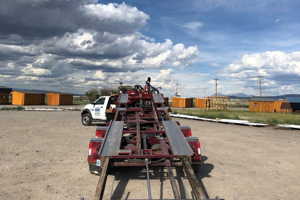 A white tow truck is parked on a gravel lot under a cloudy sky. Attached is a large, red tilt-bed tow mechanism. Several wooden cabin-like structures are in the background.