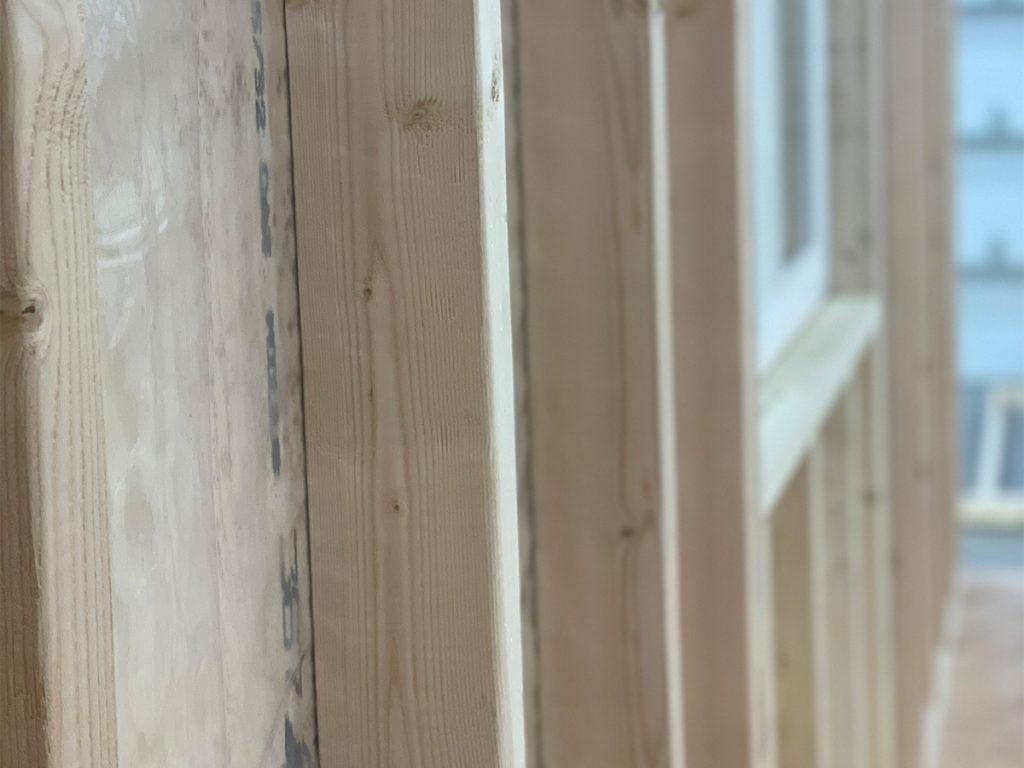 Close-up of wooden beams forming a partially constructed wall, with light filtering through the gaps. The beams have a smooth, natural finish, highlighting the texture of the wood grain.