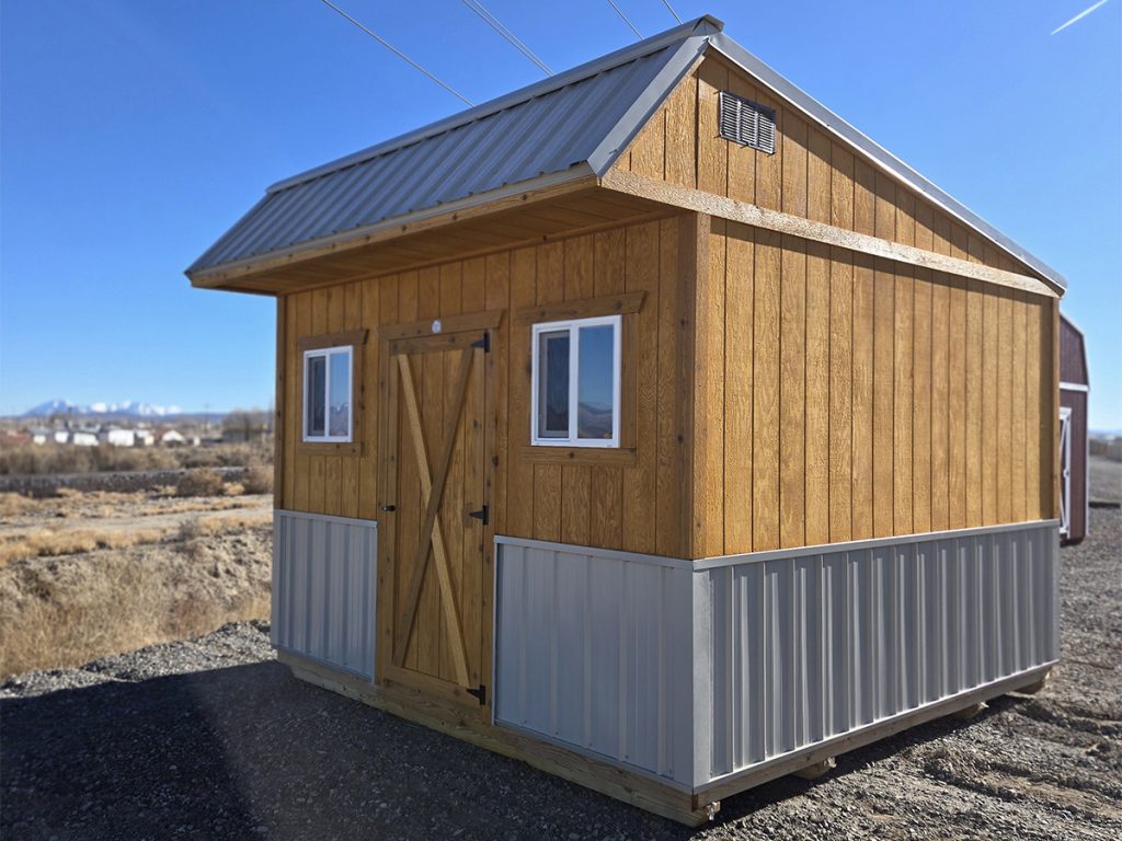 A wooden Saltbox shed with a metal roof sits on a gravel lot. It has a barn-style door and two small windows. The background features a clear blue sky and distant mountains.