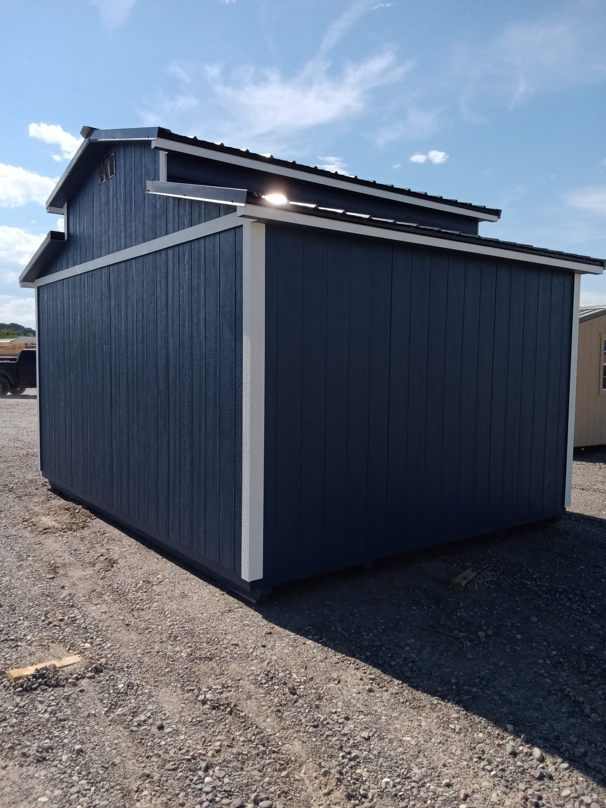 The All American Shed – 2181, painted blue with white trim and a vented roof, stands on a gravel lot beneath a sunny sky with scattered clouds. Another building appears in the background to the right.