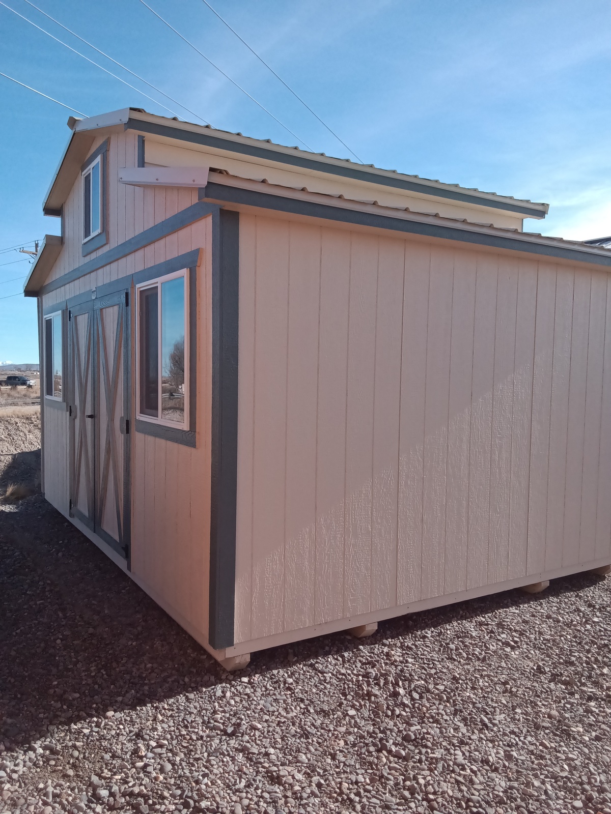 An All American Shed - 2315, beige with green trim and double barn doors, sits on gravel under a blue sky. The shed features windows and a lofted roof, providing extra storage space.