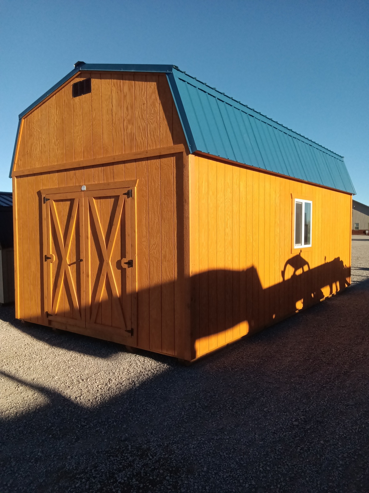 The Lofted Dutch Barn - 2419 features double barn-style doors, a blue metal roof, and a white-framed window on the right wall. It is set on gravel and illuminated by sunlight, which casts pronounced shadows on one side.