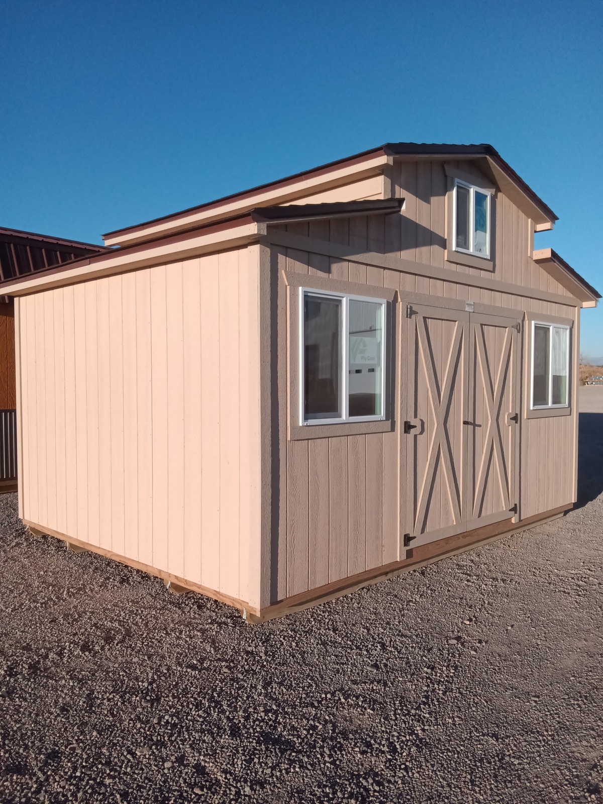 An All American Shed - 2283 in light brown and tan with double doors and two windows stands on gravel under a clear blue sky.