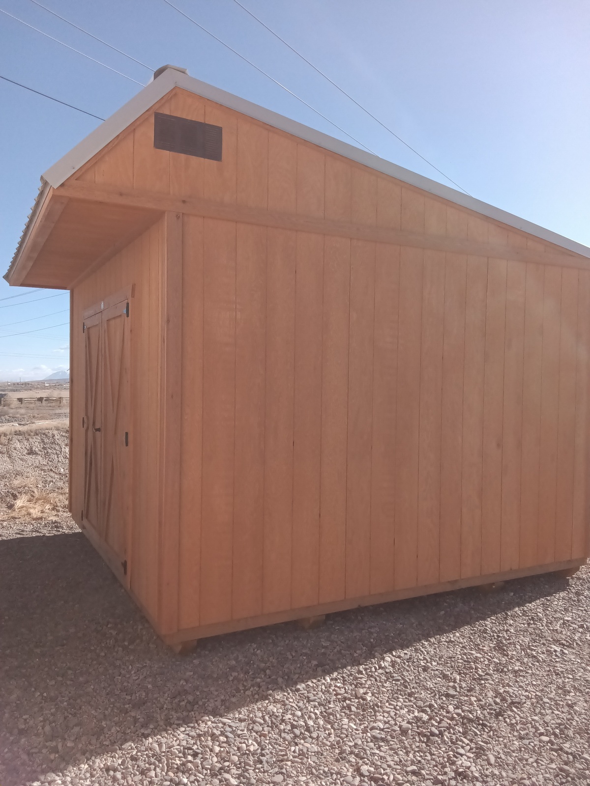 The Saltbox - 2496, a light brown wooden shed with a slanted roof and double doors, is situated on a gravel surface in a sunny area with mountains visible in the distance.