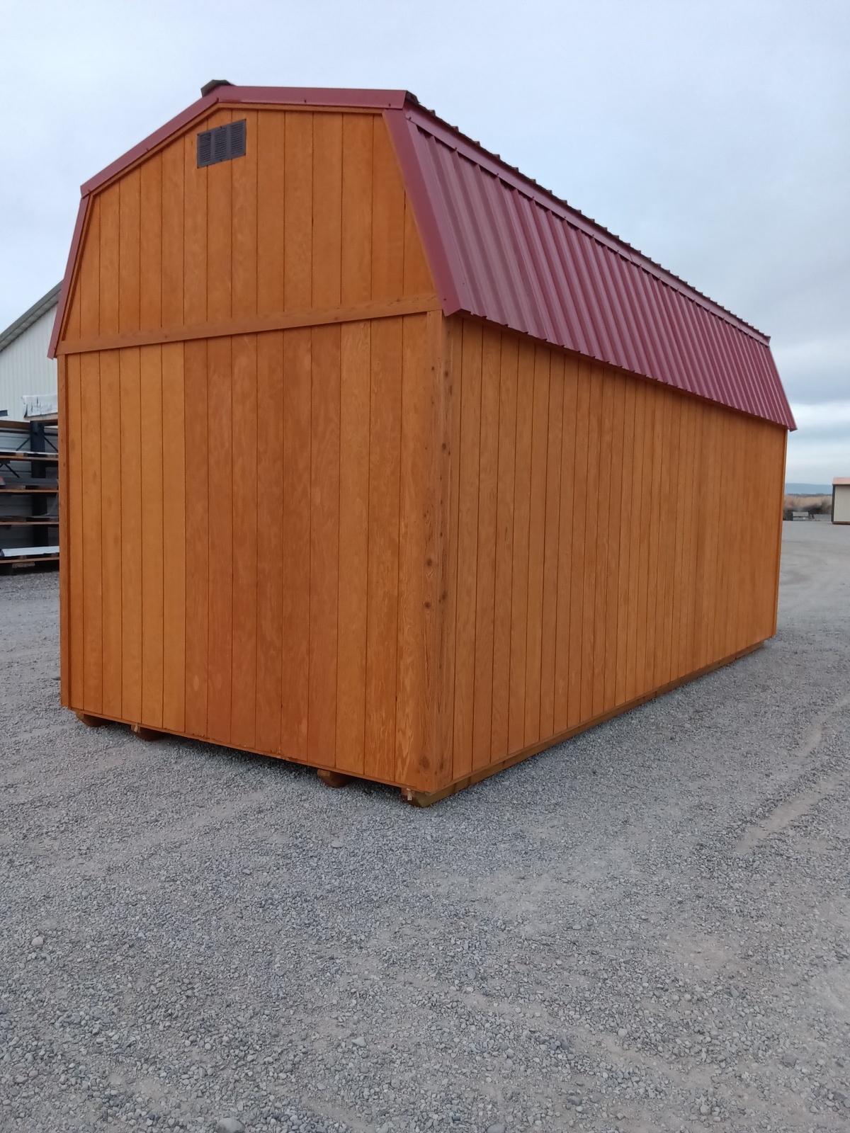 A Lofted Dutch Barn - 2493 storage shed with vertical panel siding and a red metal gambrel roof sits on a gravel lot under a cloudy sky, positioned with one rectangular side facing the camera.