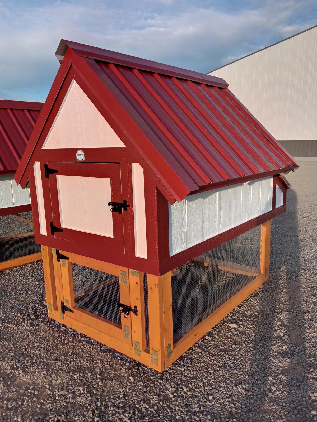 The Chicken Coop - 2445 is a compact red and white coop with a sloped metal roof, mesh lower section, and latched doors, set on outdoor gravel.