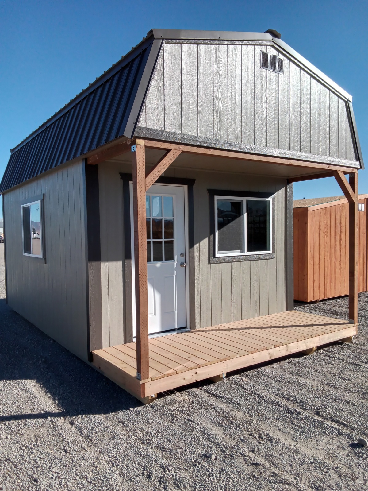 A Lofted Dutch Cabin - 2499, small and gray with a black metal roof, white door, and front porch, stands on a gravel lot under a clear blue sky with another wooden building visible in the background.