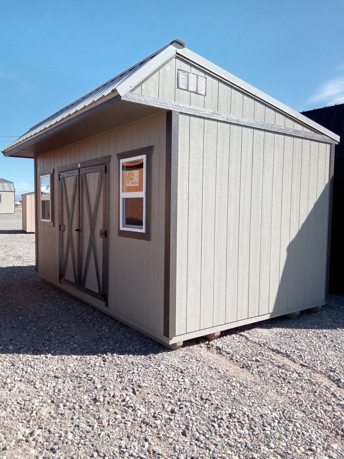 The Saltbox - 2563, a light gray wooden shed with double barn-style doors and two windows, stands on a gravel lot beneath a clear blue sky.