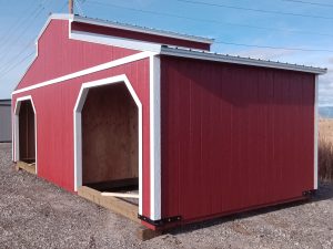 A red Westerner - 2414 barn-style shed with white trim sits on gravel under a blue sky. It features two large open doorways, a sloped metal roof, and power lines in the background.