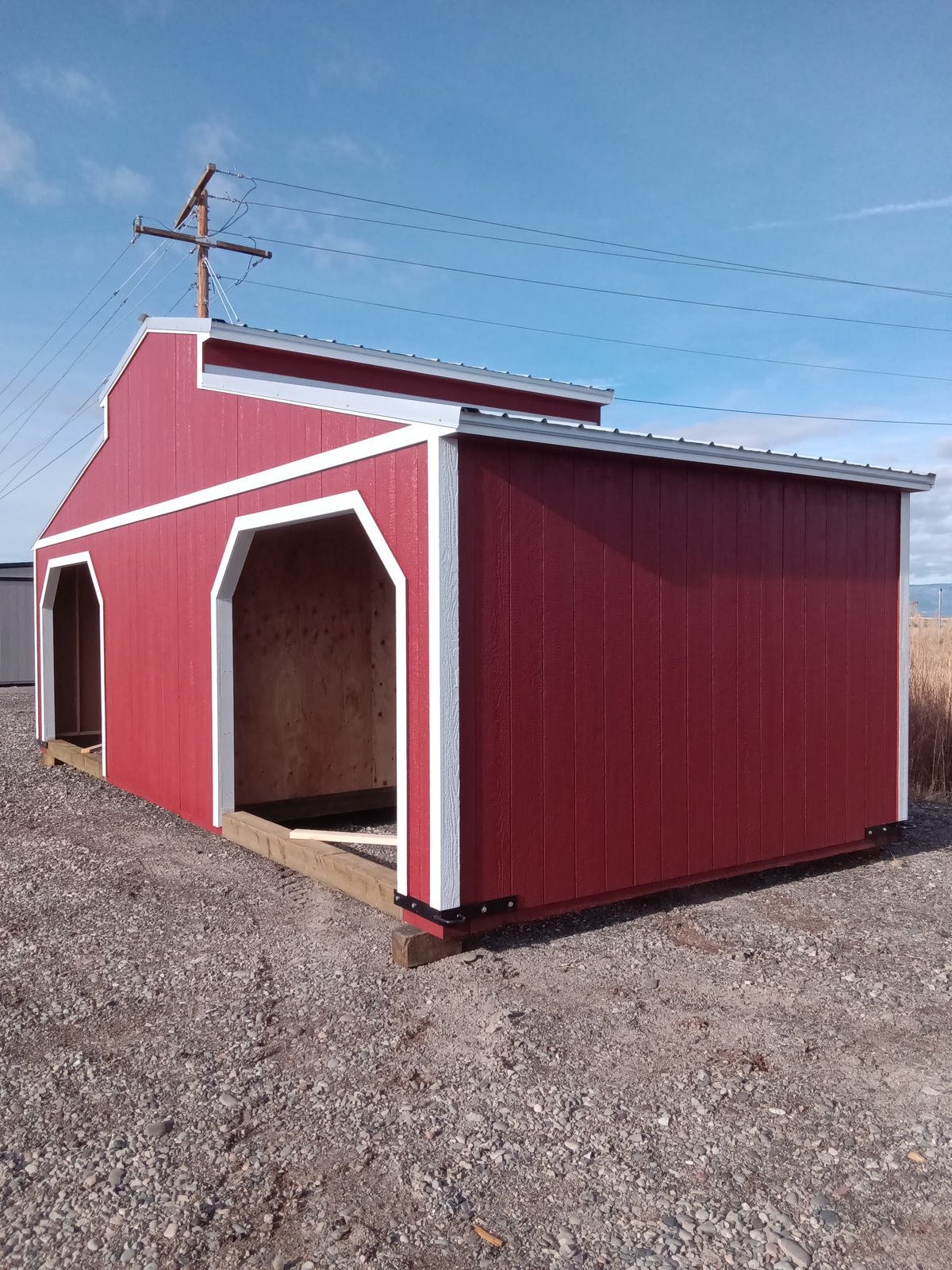 A red Westerner - 2414 barn-style shed with white trim sits on gravel under a blue sky. It features two large open doorways, a sloped metal roof, and power lines in the background.