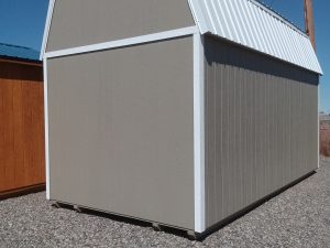 A Lofted Dutch Barn - 2570 with tan siding, white trim, and a metal roof sits on a gravel lot under a clear blue sky, with nearby power lines and a wooden post.
