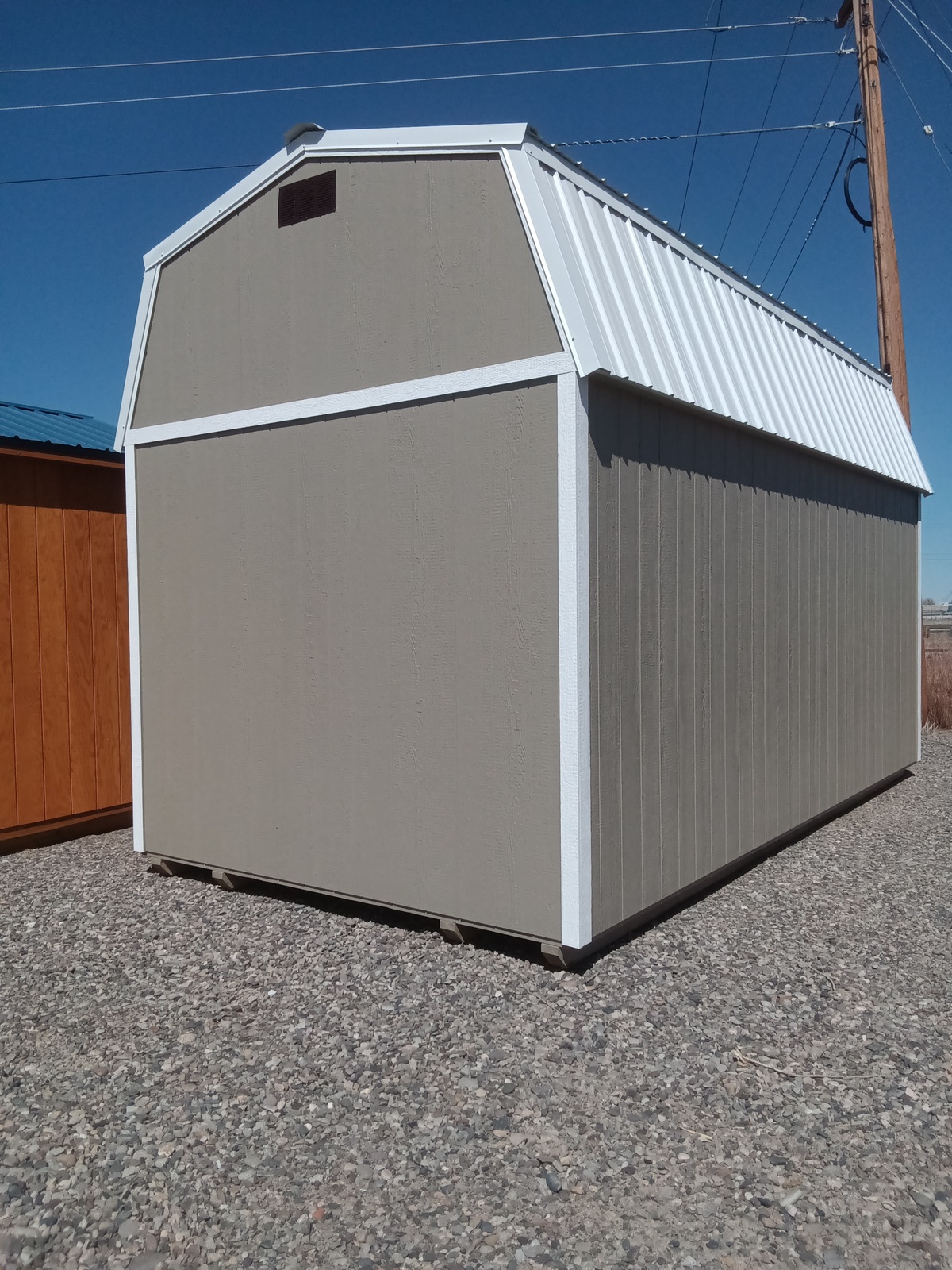 A Lofted Dutch Barn - 2570 with tan siding, white trim, and a metal roof sits on a gravel lot under a clear blue sky, with nearby power lines and a wooden post.