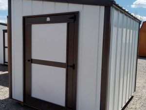 The Lean-To - 2671 is a compact white metal storage shed with a dark brown frame and door, shown outdoors on gravel under a partly cloudy sky.