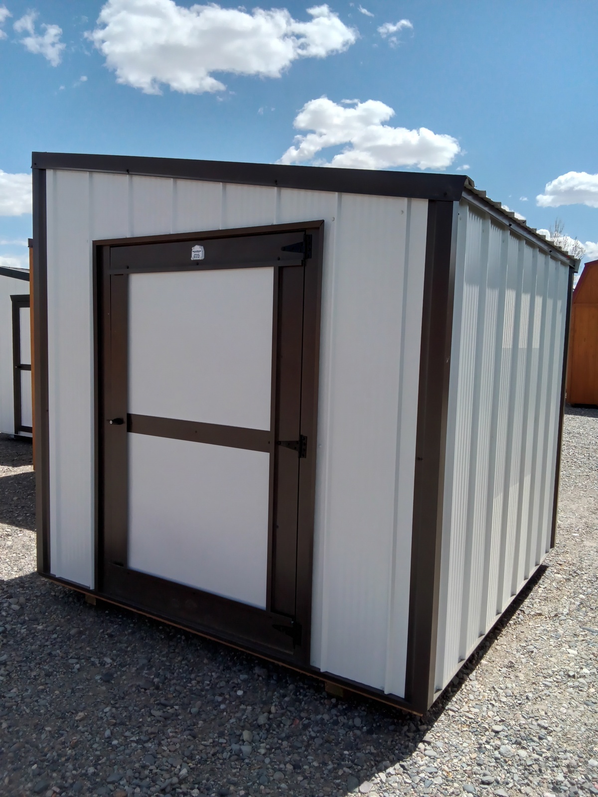 The Lean-To - 2671 is a compact white metal storage shed with a dark brown frame and door, shown outdoors on gravel under a partly cloudy sky.