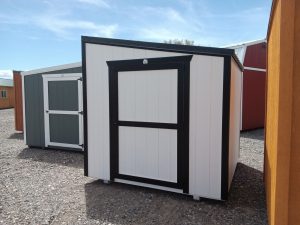 A Lean-To - 2669, a small white shed with black trim and a black-framed door, stands on gravel. In the background, there’s a green and white shed and a red shed under a blue sky.
