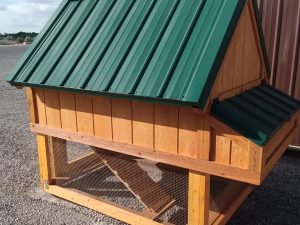 The Chicken Coop - 2668 features a wooden structure with a green metal roof, wire mesh on the lower section, and a small gable extension. It stands on gravel with a partly cloudy sky in the background.