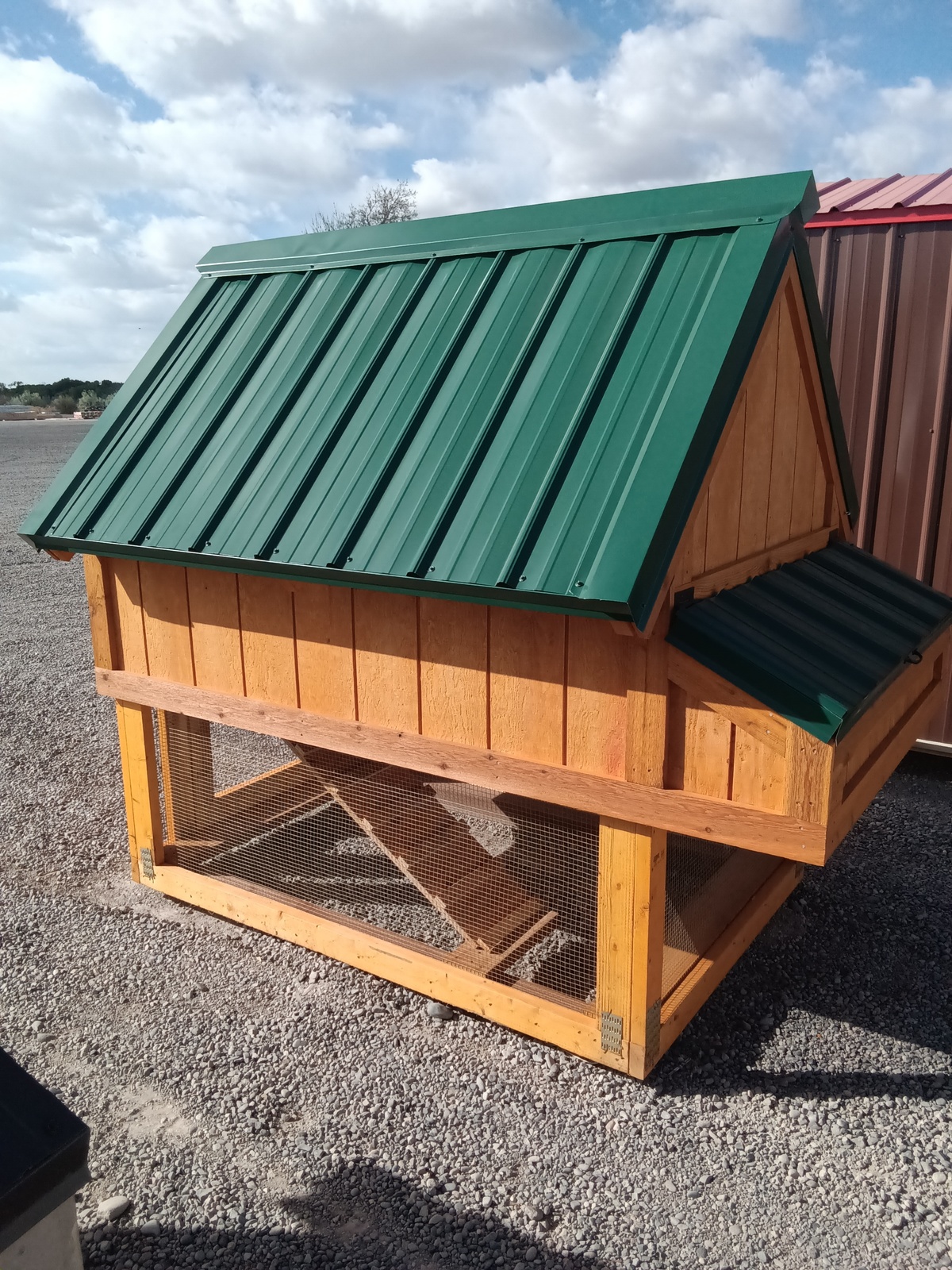 The Chicken Coop - 2668 features a wooden structure with a green metal roof, wire mesh on the lower section, and a small gable extension. It stands on gravel with a partly cloudy sky in the background.