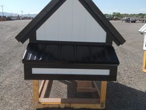 The Chicken Coop - 2665 is a small wooden coop with black and white roof and siding, set on a wire mesh base outdoors on gravel under a clear blue sky.