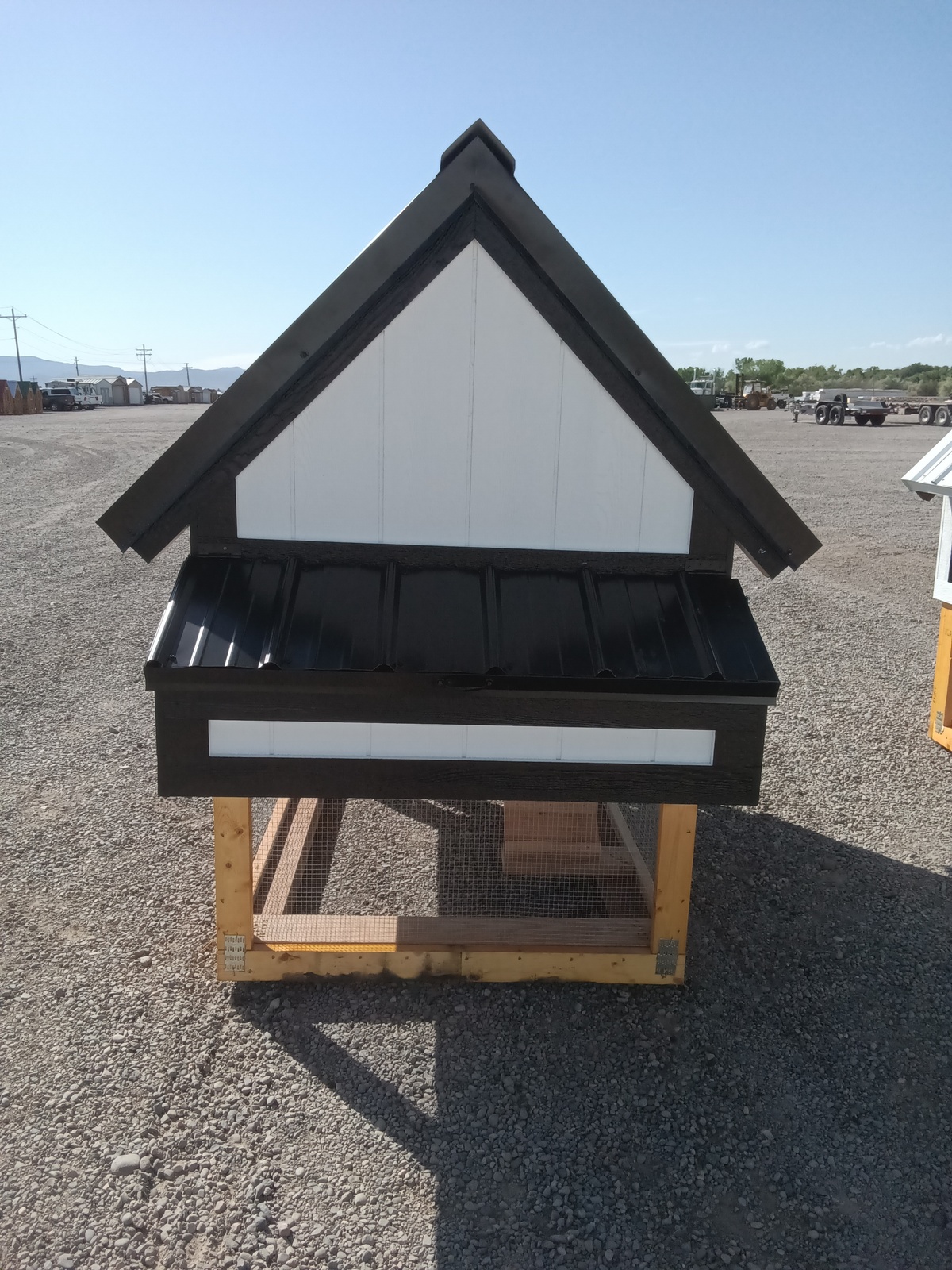 The Chicken Coop - 2665 is a small wooden coop with black and white roof and siding, set on a wire mesh base outdoors on gravel under a clear blue sky.