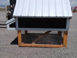 The Chicken Coop - 2666, featuring a white metal roof, black walls, and a wooden frame, stands outdoors on gravel. A forklift appears on the left with clear skies in the background.