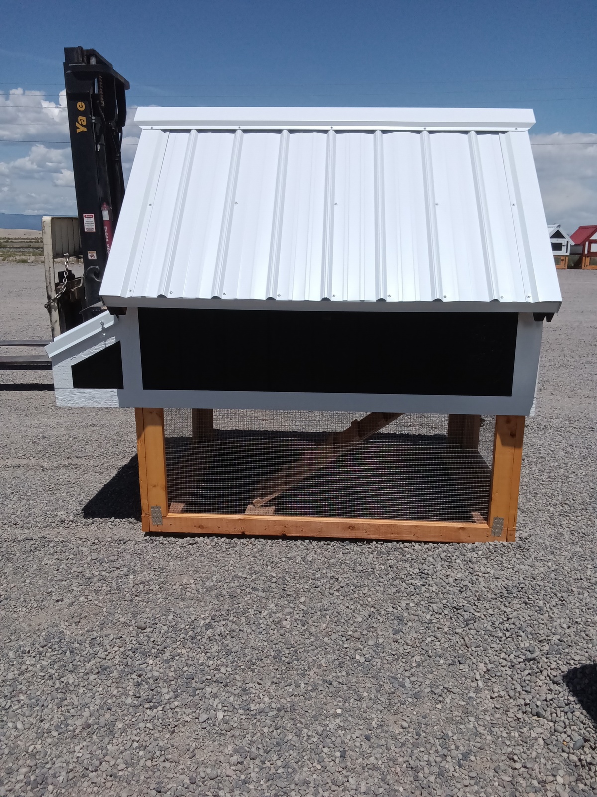 The Chicken Coop - 2666, featuring a white metal roof, black walls, and a wooden frame, stands outdoors on gravel. A forklift appears on the left with clear skies in the background.