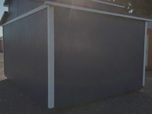 The All American Shed - 2805 with white trim sits on gravel under bright sunlight, casting long shadows. Trees can be seen in the background.