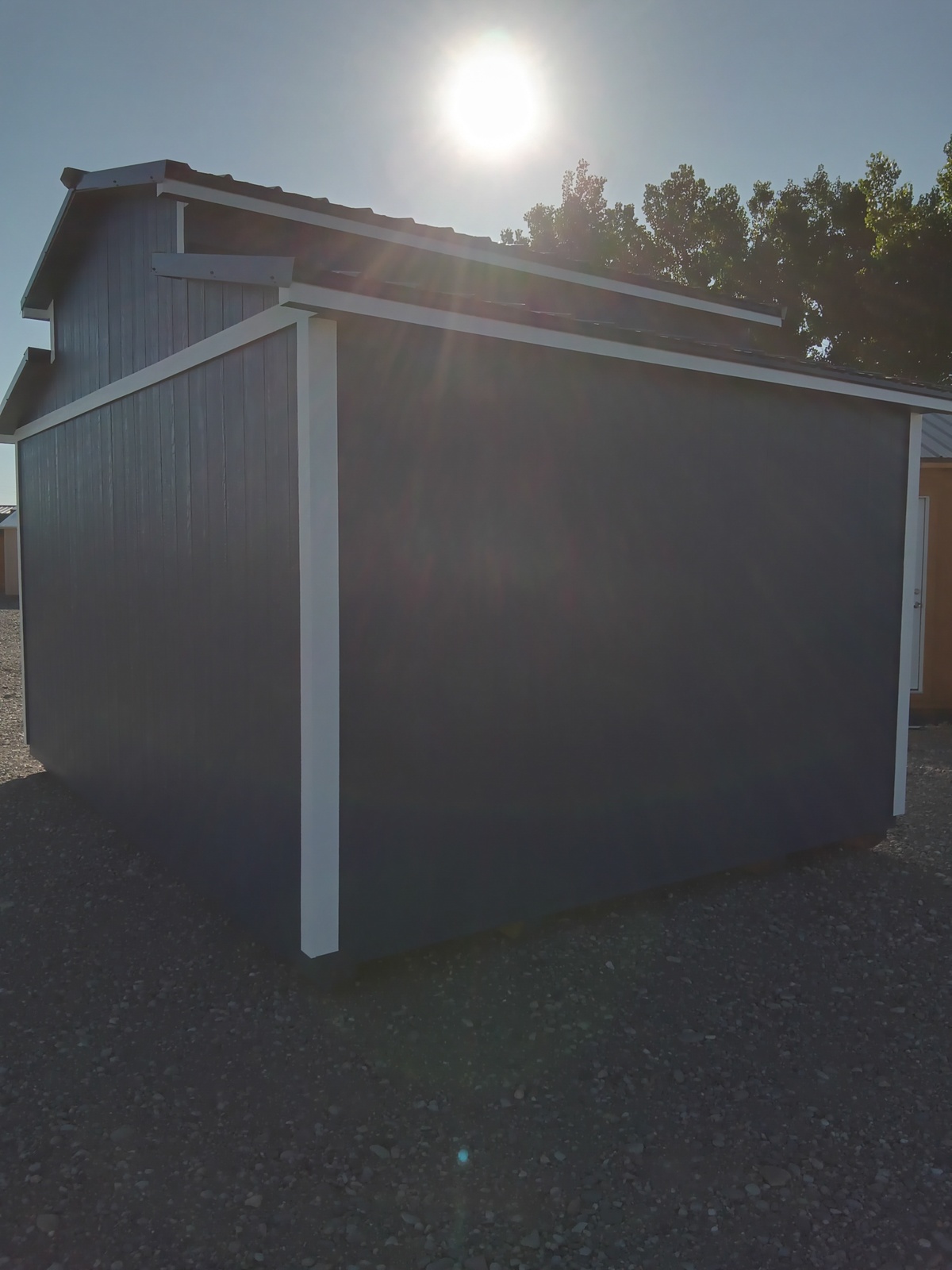 The All American Shed - 2805 with white trim sits on gravel under bright sunlight, casting long shadows. Trees can be seen in the background.