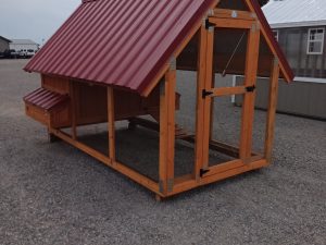 The Chicken House - 2879 is a wooden coop with a red metal roof, a side nesting box, and a wire-enclosed area below. It sits on gravel with buildings visible in the background beneath cloudy skies.