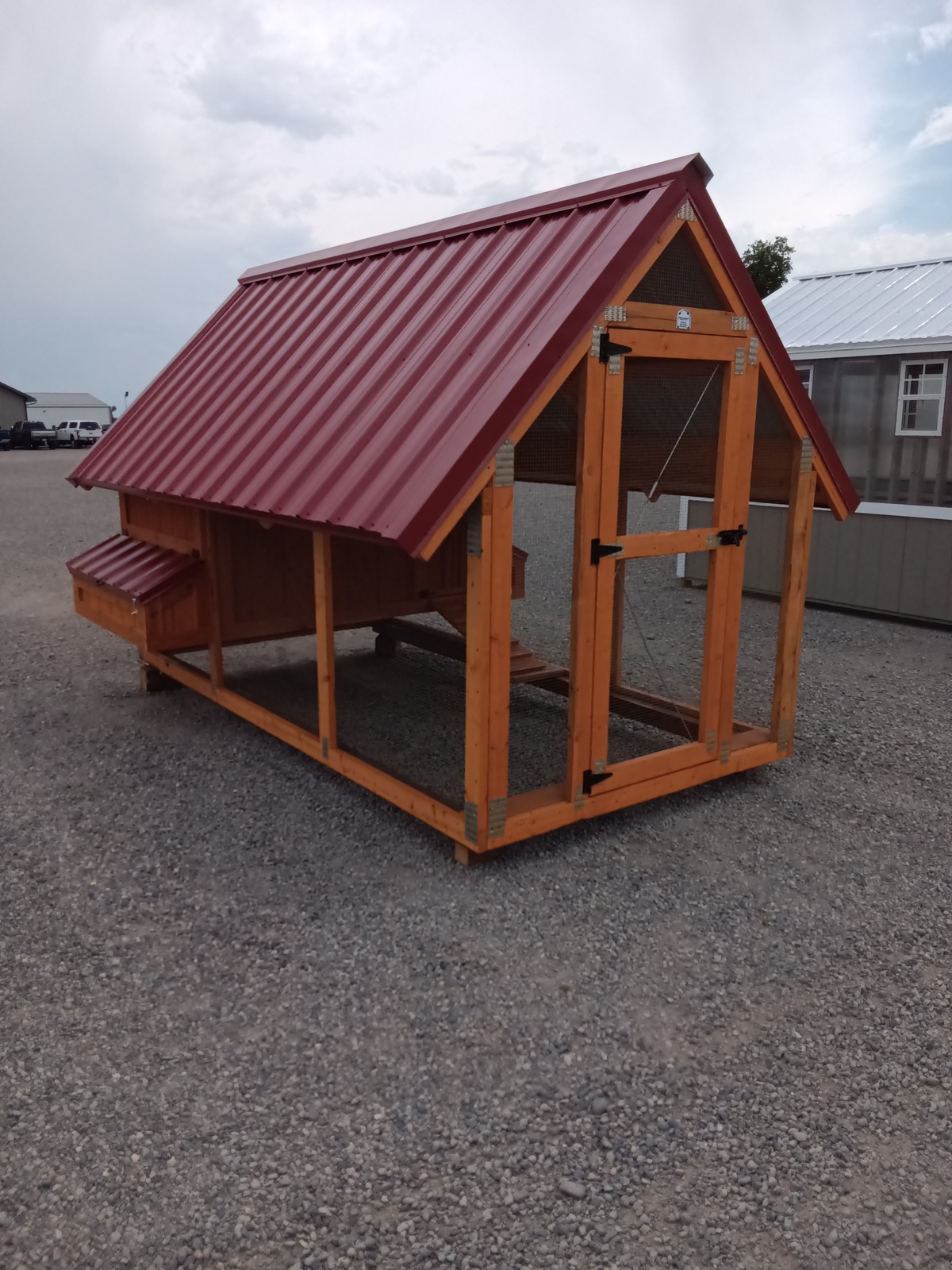 The Chicken House - 2879 is a wooden coop with a red metal roof, a side nesting box, and a wire-enclosed area below. It sits on gravel with buildings visible in the background beneath cloudy skies.