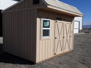 A Saltbox - 1152 beige wooden shed with double doors and a small window sits on gravel near a larger white building under a clear blue sky, its unique design adding charm to the peaceful setting.