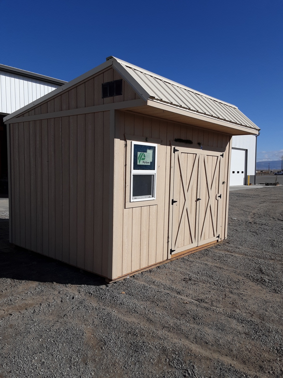 A Saltbox - 1152 beige wooden shed with double doors and a small window sits on gravel near a larger white building under a clear blue sky, its unique design adding charm to the peaceful setting.