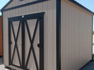 A tan Classic Shed - 2910 with black trim and double doors featuring an "X" design sits on gravel under a clear blue sky.