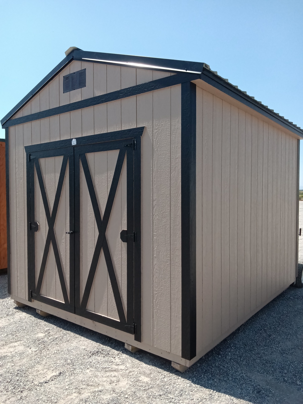 A tan Classic Shed - 2910 with black trim and double doors featuring an "X" design sits on gravel under a clear blue sky.