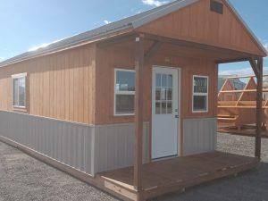 The Mountain Cabin - 3017 is a small wooden cabin featuring a front porch, white door, and multiple windows, set on gravel beneath a clear, sunny sky. Additional Mountain Cabin - 3017 units are visible in the background.