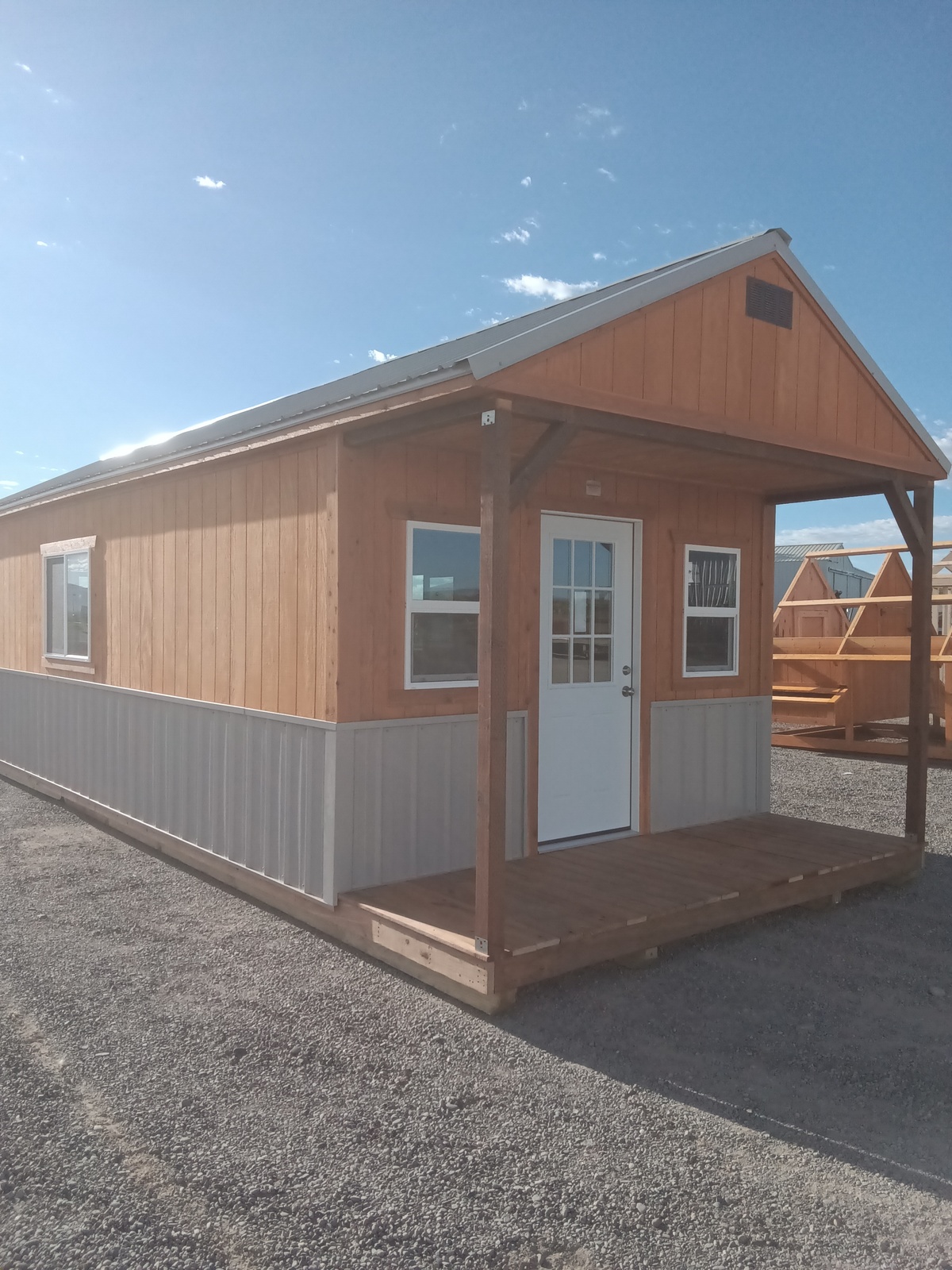 The Mountain Cabin - 3017 is a small wooden cabin featuring a front porch, white door, and multiple windows, set on gravel beneath a clear, sunny sky. Additional Mountain Cabin - 3017 units are visible in the background.