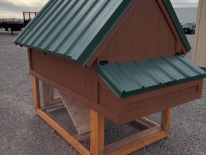 The Chicken Coop - 3027 is a small brown wooden coop with a green metal roof, raised design, mesh bottom, and sloping roof panels. It stands on gravel outdoors under a cloudy sky.
