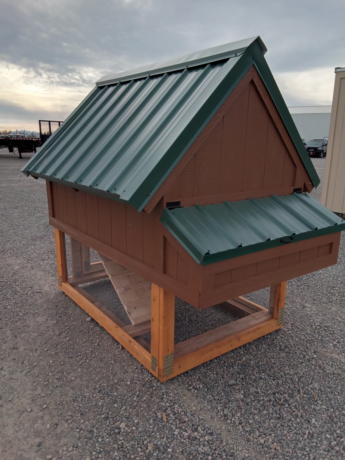 The Chicken Coop - 3027 is a small brown wooden coop with a green metal roof, raised design, mesh bottom, and sloping roof panels. It stands on gravel outdoors under a cloudy sky.