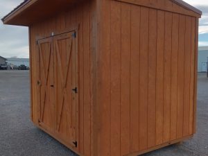 The Saltbox - 3150, a compact wooden storage shed with double doors and a red slanted roof, stands on a gravel lot beneath a cloudy sky.