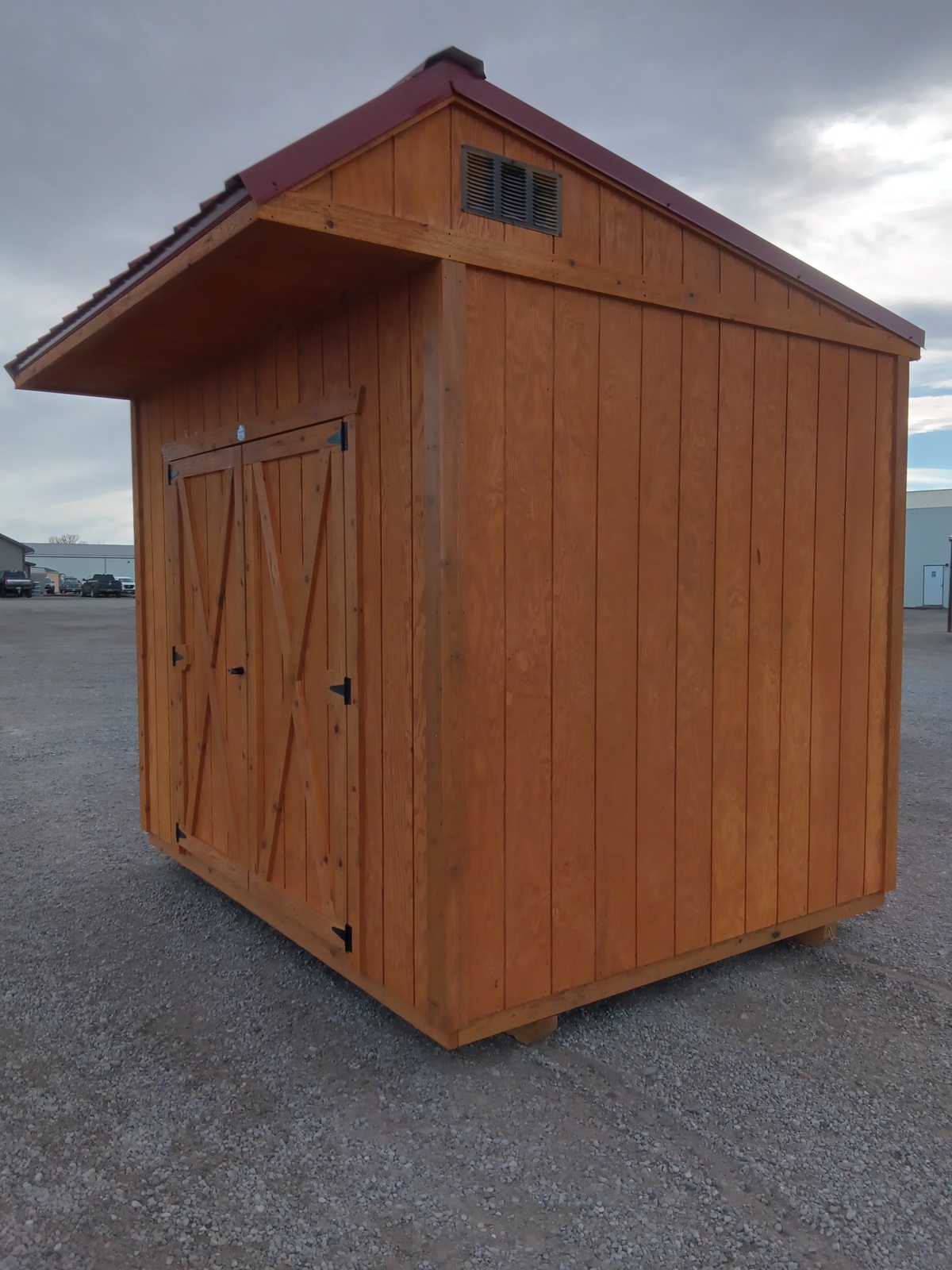 The Saltbox - 3150, a compact wooden storage shed with double doors and a red slanted roof, stands on a gravel lot beneath a cloudy sky.
